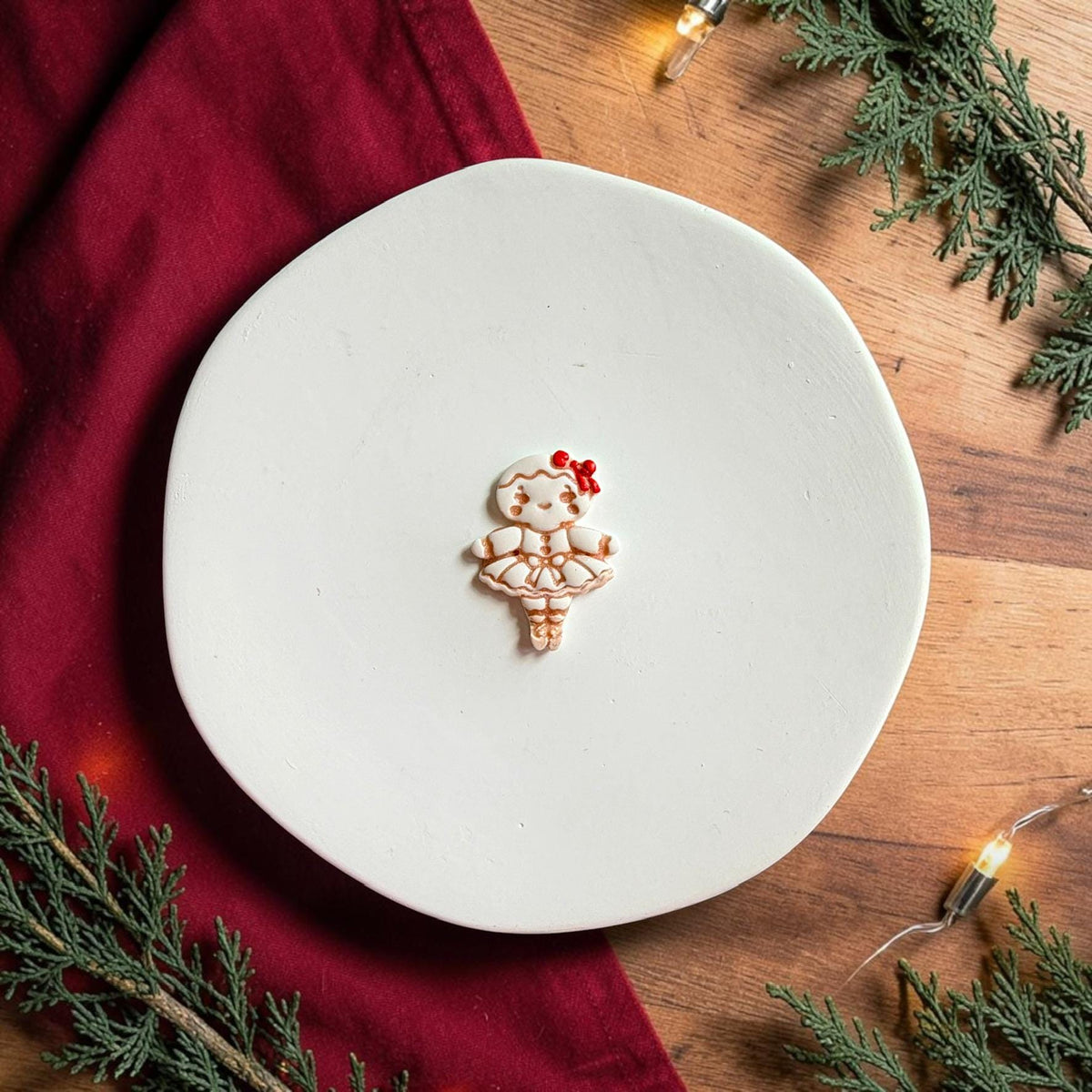 Finished white gingerbread ballerina clay piece with brown decorative details and a small red bow centered on a white plate with red fabric and Christmas greenery.