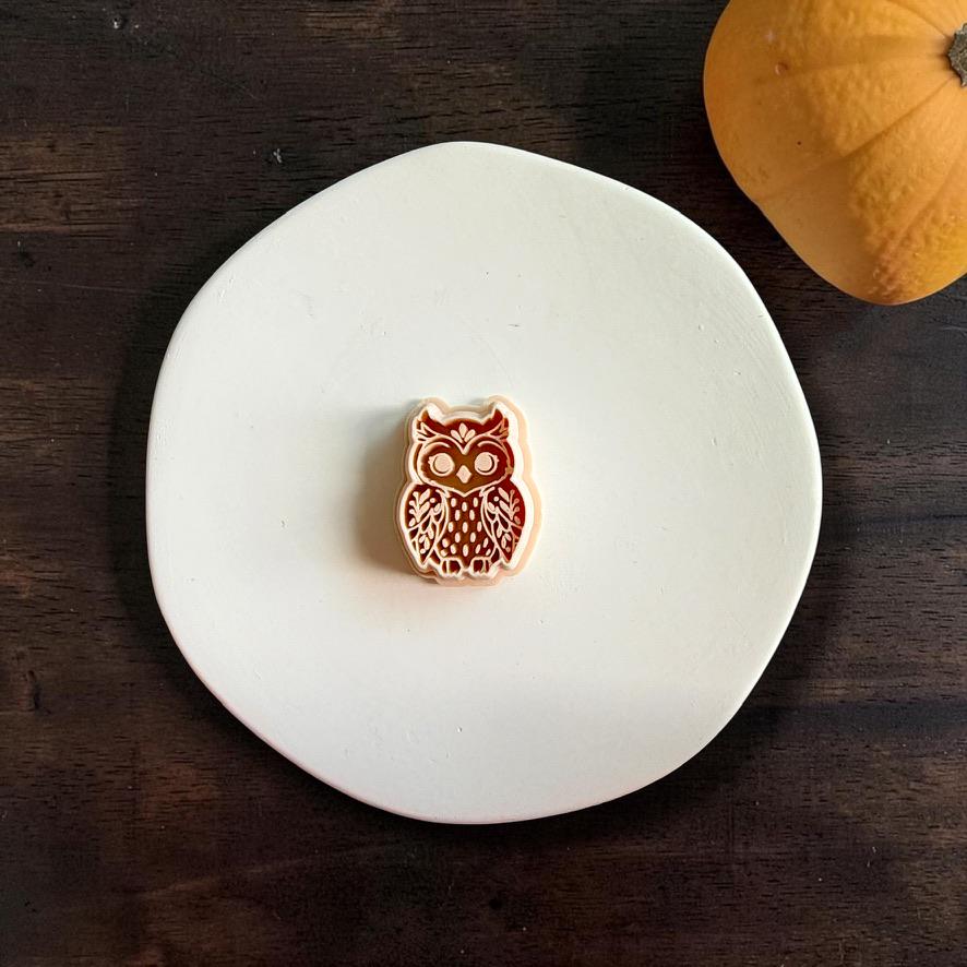 Ornate owl polymer clay cutter centered on a white plate, showing the detailed eyes, wings, feathers, and decorative body pattern, with a small pumpkin in the background.
