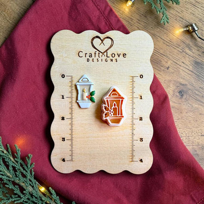 Finished white Christmas lantern clay piece with a candle and green holly leaves beside the matching polymer clay cutter on a wooden Craft Love Designs sizing board with red fabric, greenery, and warm holiday lights.