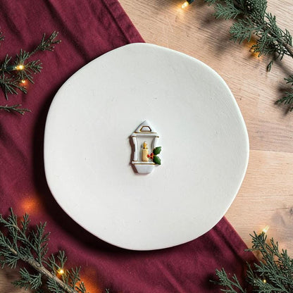 Finished white Christmas lantern clay piece centered on a white plate, featuring a candle with a yellow flame, green holly leaves, and red berries against a festive red fabric and evergreen background.
Photo 3: