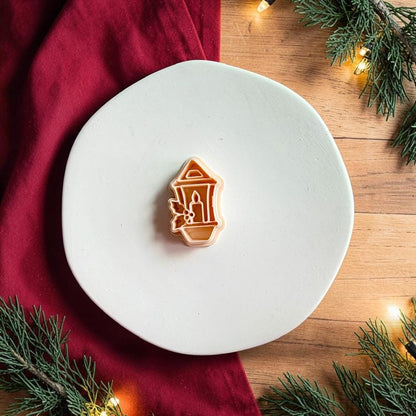 Christmas lantern polymer clay cutter centered on a white plate, showing the lantern outline, interior candle, and holly detail with red holiday fabric and evergreen branches nearby.