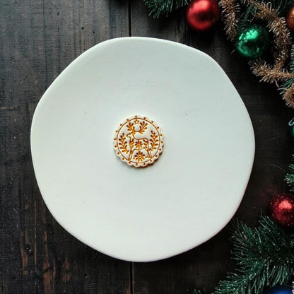 Small white and metallic gold folk art reindeer design centered on a white round surface, showing the deer, antlers, flowers, botanical branches, dotted scalloped border, and tiny star surrounded by festive Christmas decor.