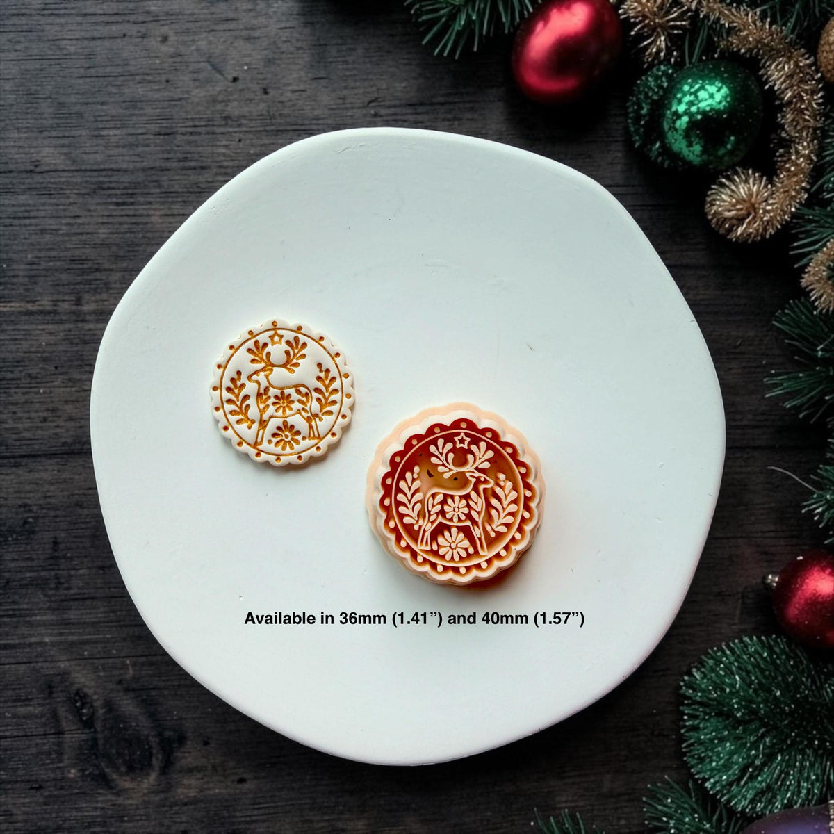 White and gold 36mm reindeer design beside the larger red and white 40mm version on a white surface. Both show the circular botanical folk art deer pattern. Text states available in 36mm (1.41") and 40mm (1.57").