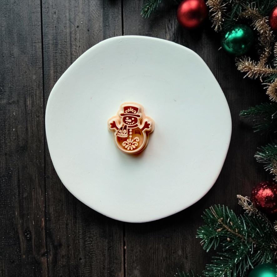 Large red and cream folk art snowman design centered on a white surface. The snowman features a decorated top hat, scarf, buttons, raised arms, smiling face, and ornamental floral motif, surrounded by festive Christmas decorations.
