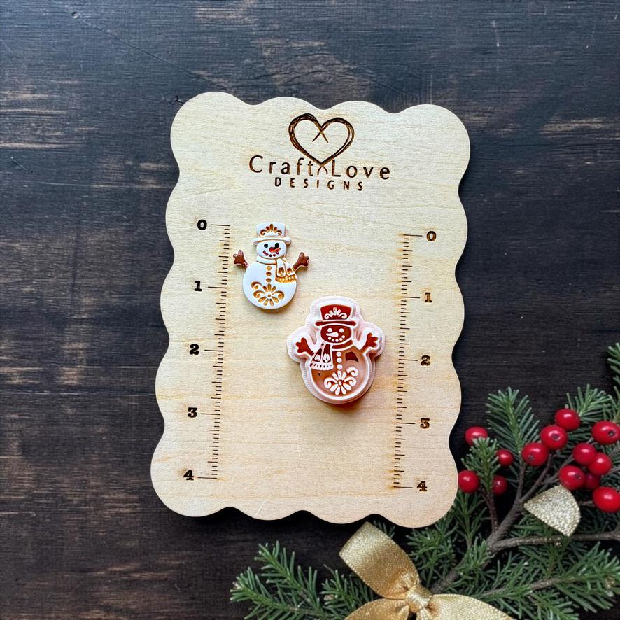 Two sizes of a decorative folk art snowman displayed on a Craft Love Designs wooden measuring board. The snowman has a top hat, scarf, outstretched arms, buttons, and floral detailing, with Christmas greenery, berries, and a gold bow nearby.