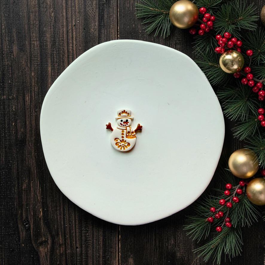 Small white, gold, red, and black finished snowman centered on a white surface with evergreen branches, red berries, and gold Christmas ornaments along the edge.