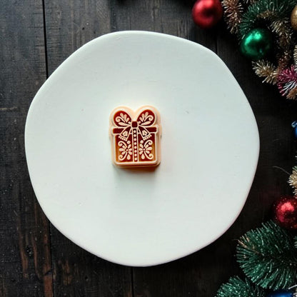 Large red and cream Christmas present design centered on a white surface. The wrapped gift features an oversized bow, central ribbon, dotted accents, and symmetrical folk art floral details, with Christmas greenery and ornaments along the edge.