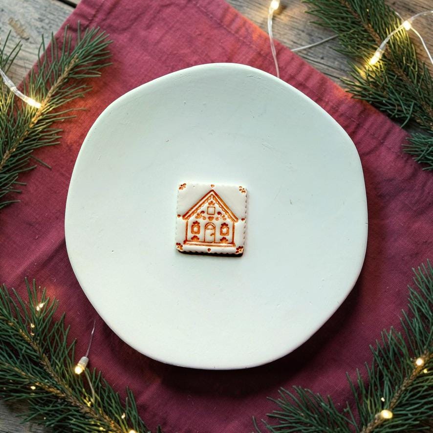 Small white gingerbread house design centered on a white plate over a red cloth, surrounded by evergreen branches and warm white Christmas lights.