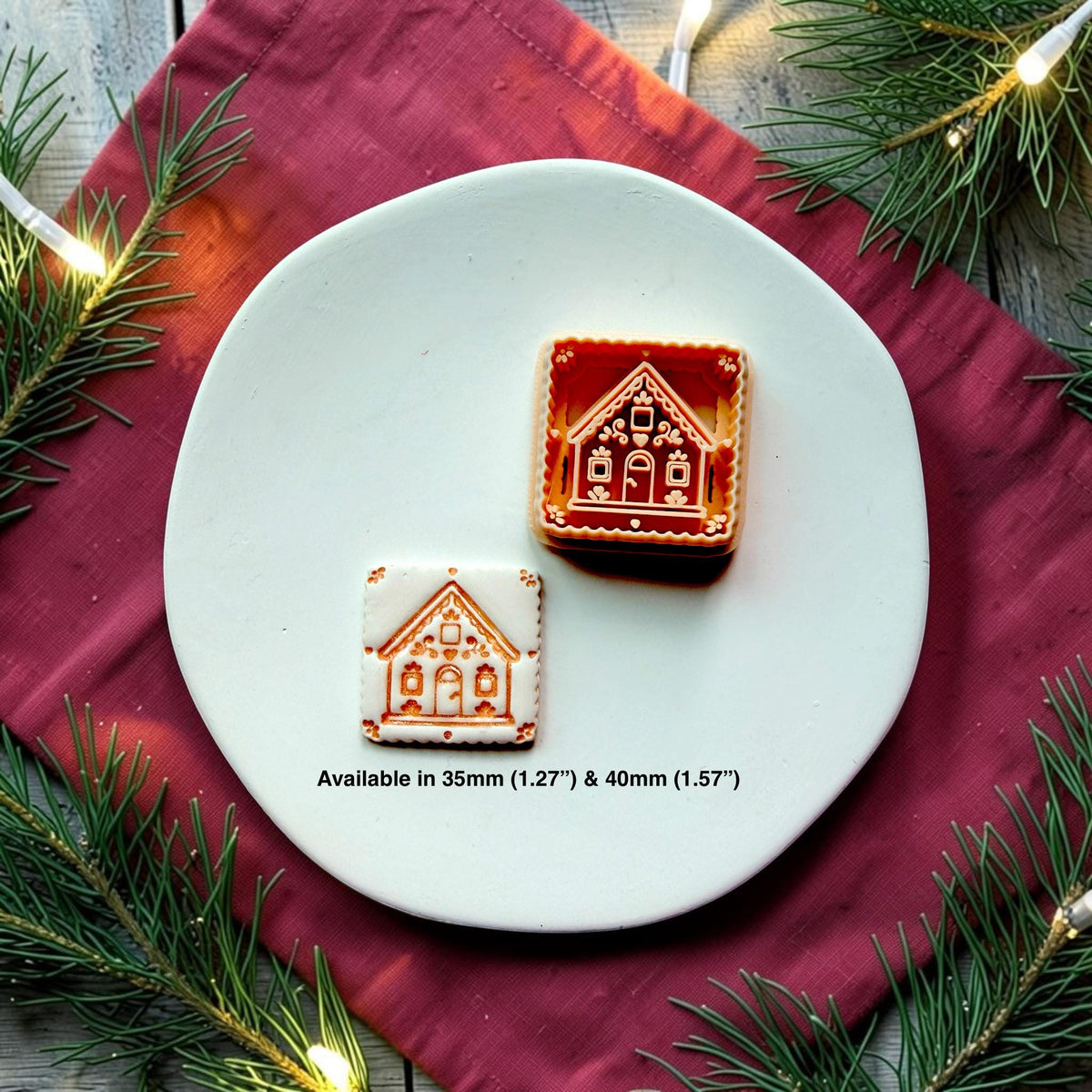 Small white and brown gingerbread house beside a larger brown and cream version on a white plate surrounded by evergreen branches and glowing Christmas lights. Text shows available sizes of 35mm (1.27") and 40mm (1.57")