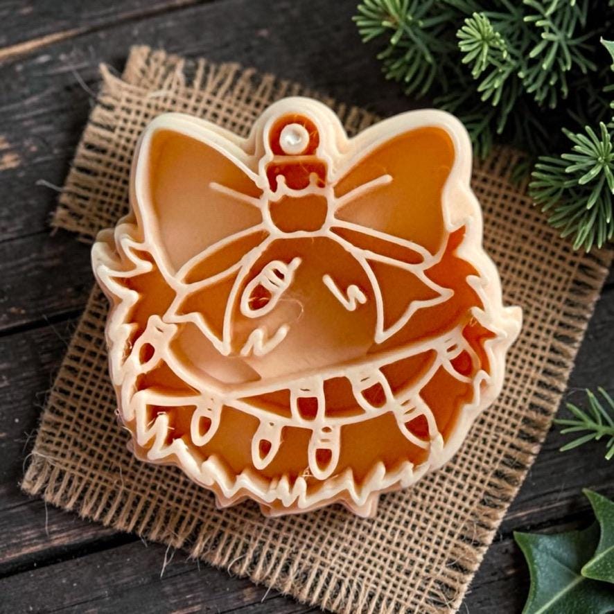 Close-up of the tan clay cutter showing the ornament shape with an oversized bow, shaggy edging, hanging Christmas lights, and detailed interior lines, displayed on burlap with evergreen accents.