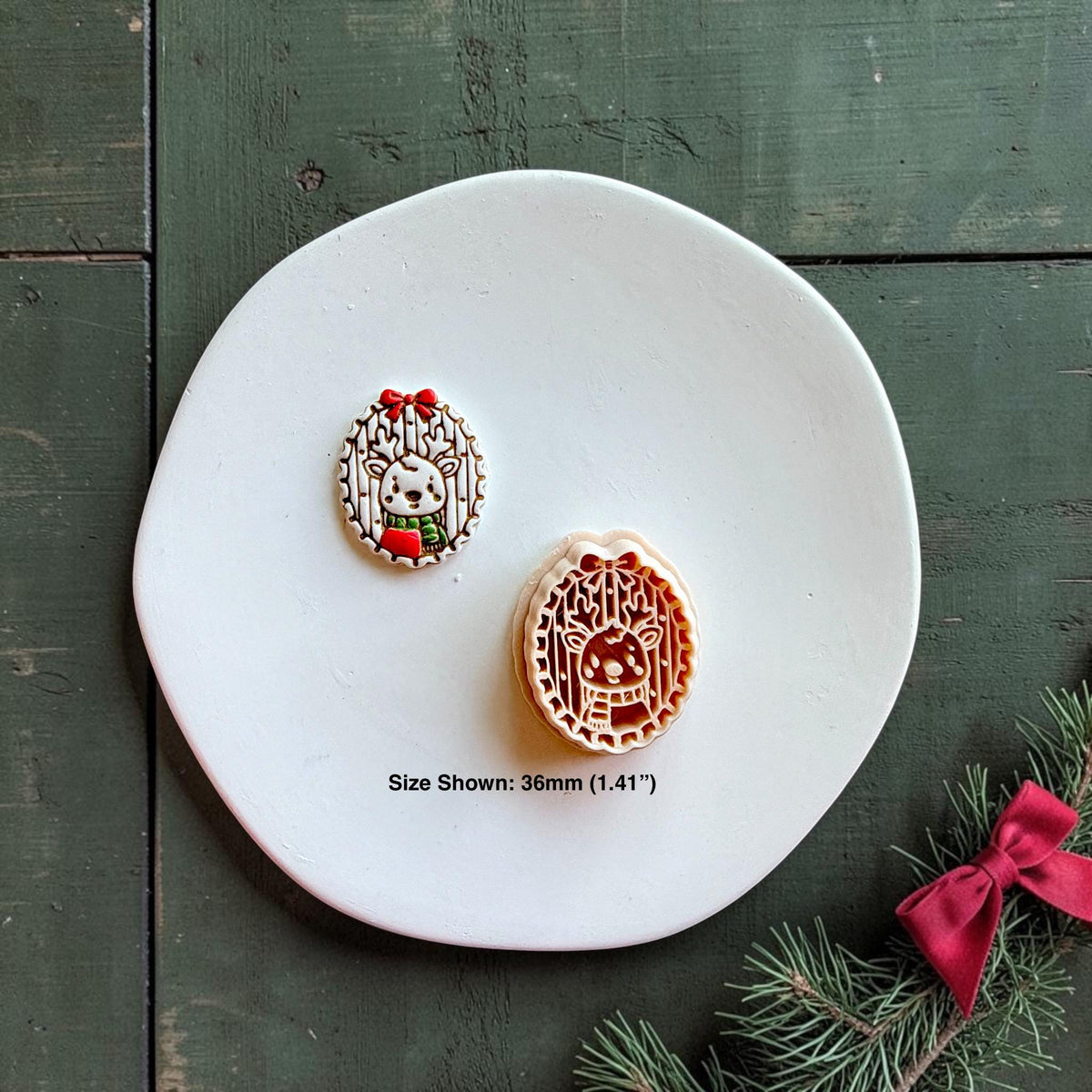 Finished reindeer design and matching clay cutter displayed together on a white plate over dark green wood, with evergreen branches and a red bow; text indicates the size shown is 36mm (1.41")