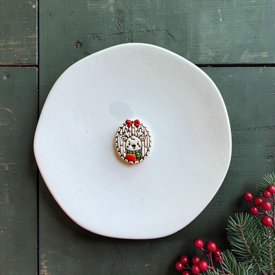 Finished Christmas reindeer piece centered on a white plate, featuring a small deer with antlers and scarf inside an oval woodland frame topped with a red bow, with evergreen and red berries nearby