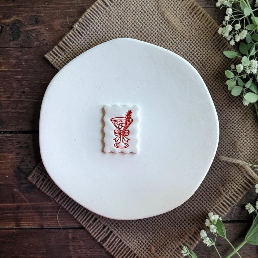 Finished white scalloped rectangle clay piece centered on a white plate, featuring a red martini glass with a bow and decorative leafy sprig against a rustic wood and burlap background.