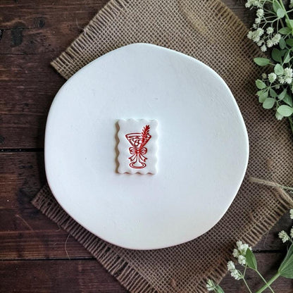 Finished white scalloped rectangle clay piece centered on a white plate, featuring a red martini glass with a bow and decorative leafy sprig against a rustic wood and burlap background.
