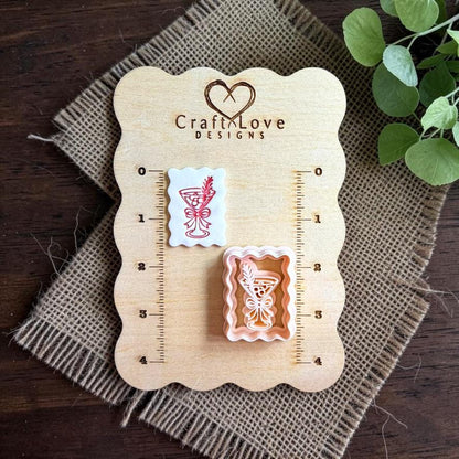 Finished white scalloped rectangular clay piece with a red martini glass, bow, and leafy sprig beside the matching polymer clay cutter on a wooden Craft Love Designs sizing board with burlap and greenery.