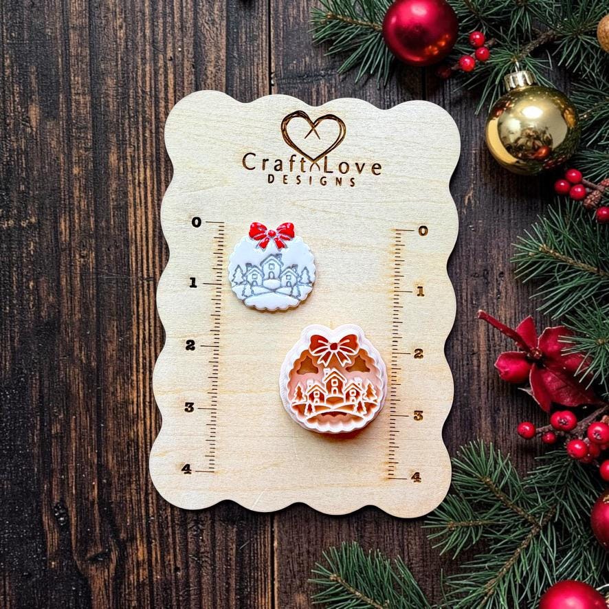 Finished white snowy Christmas village design and matching clay cutter displayed on a Craft Love Designs wooden measurement board, showing three houses, evergreen trees, snowdrifts, and a red bow, with Christmas greenery and ornaments nearby.