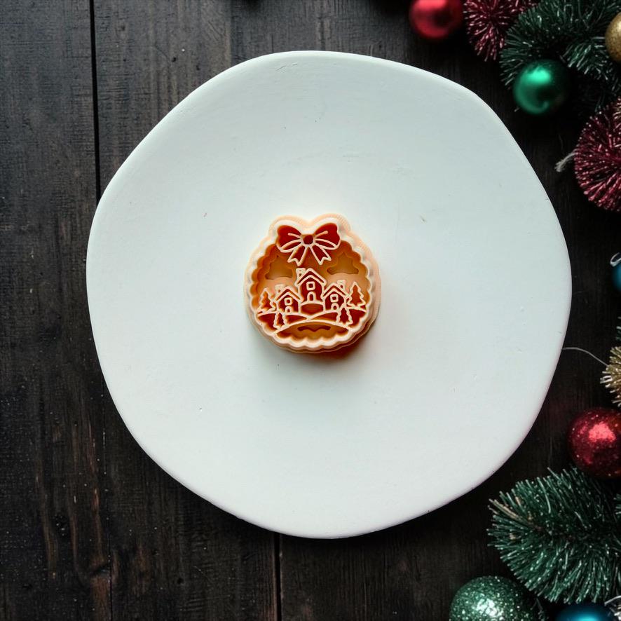 Christmas village clay cutter centered on a white plate, featuring three small houses among evergreen trees and snowy hills inside a scalloped round frame topped with a large bow.