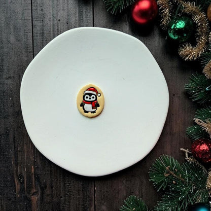 Finished Christmas penguin piece centered on a white plate, featuring a black and white penguin in a red Santa hat and scarf surrounded by festive Christmas greenery and ornaments.
