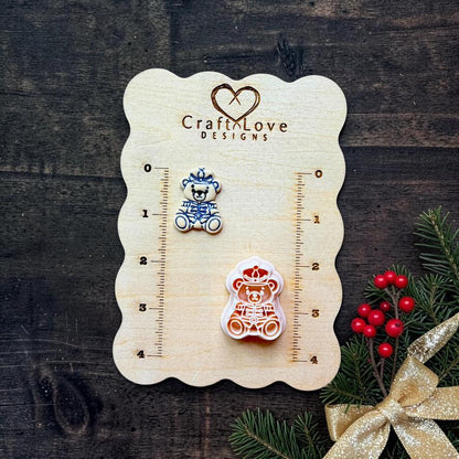 Finished teddy bear dressed as a nutcracker soldier beside the matching larger design on a Craft Love Designs wooden measurement board, surrounded by Christmas greenery, red berries, and a gold bow.
