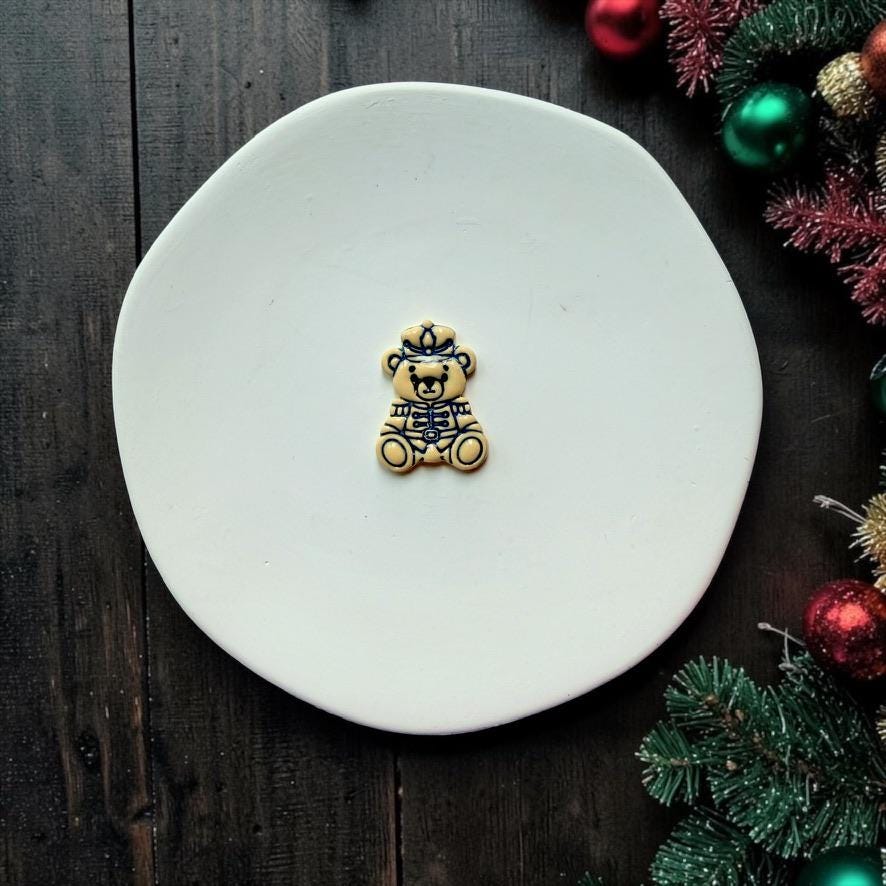 Finished teddy bear nutcracker piece centered on a white plate, showing the detailed soldier hat, jacket, belt, and seated bear shape with festive Christmas decorations along the edge.