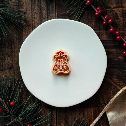 Nutcracker teddy bear design centered on a white plate, showing the seated bear wearing a decorative soldier hat and uniform, with evergreen branches, red berries, and gold ribbon around the scene.