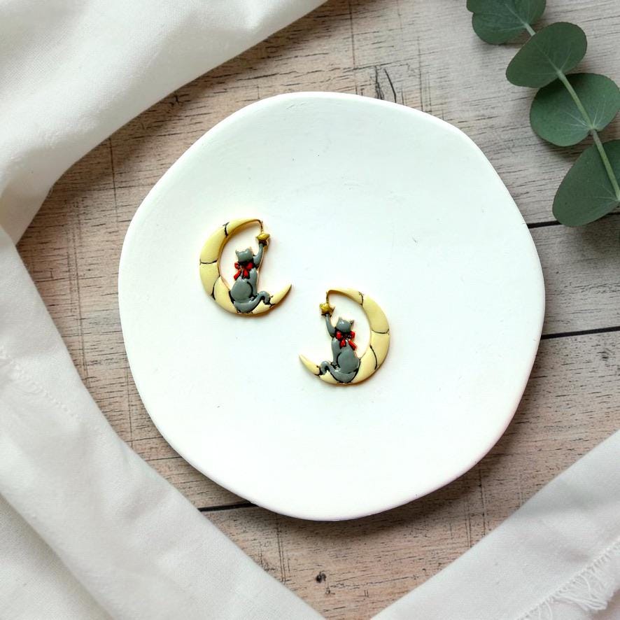 Finished mirrored pair of gray cats sitting inside pale crescent moons and reaching toward small gold stars, displayed together on a white plate.