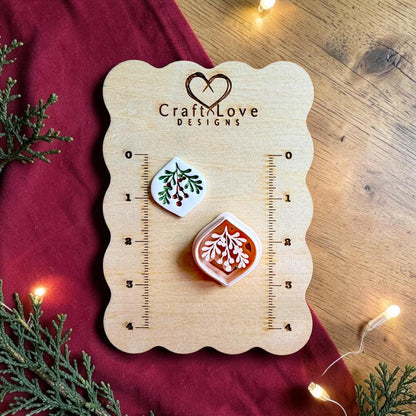 Finished white mistletoe clay piece with green leaves and red berries beside the matching polymer clay cutter on a wooden Craft Love Designs sizing board with red fabric, evergreen branches, and warm holiday lights.