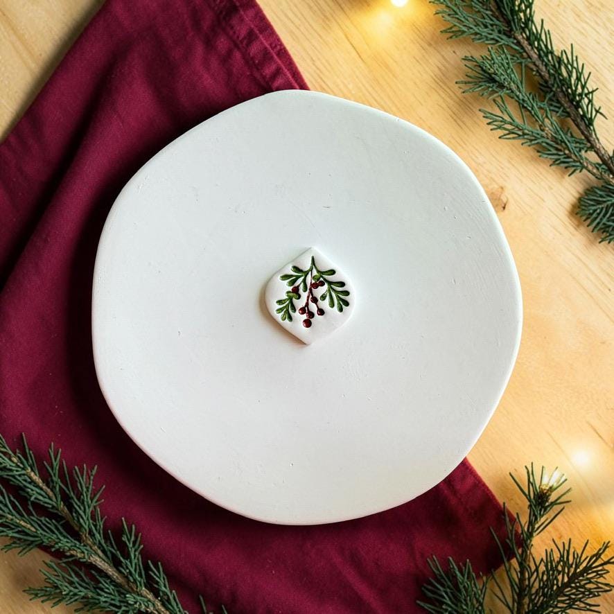 Finished white mistletoe clay piece centered on a white plate, featuring green leafy branches and red berries against a festive red fabric, wood, and evergreen background.