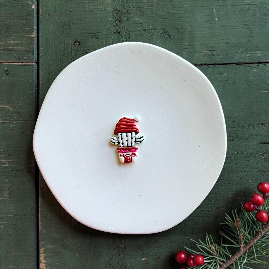 Finished Christmas cactus clay piece centered on a white plate, featuring a green and white cactus, red Santa hat, and pink-and-white decorative pot with evergreen branches and red berries nearby.