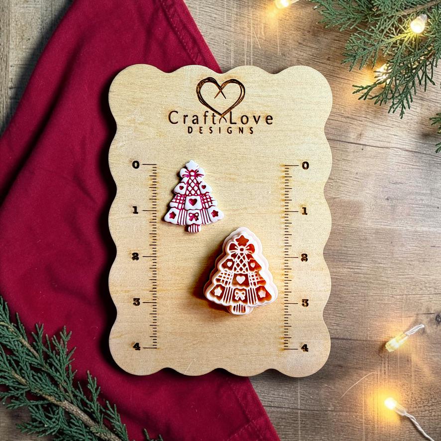 Finished white Christmas tree clay piece with dark red heart, bow, stripe, and decorative pattern details beside the matching polymer clay cutter on a wooden Craft Love Designs sizing board with red fabric, greenery, and warm lights.