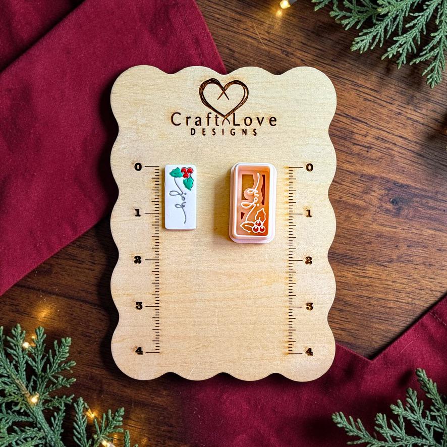 Finished white rectangular Joy clay piece with black script lettering, green holly leaves, and red berries beside the matching polymer clay cutter on a wooden Craft Love Designs sizing board with red fabric, greenery, and holiday lights.