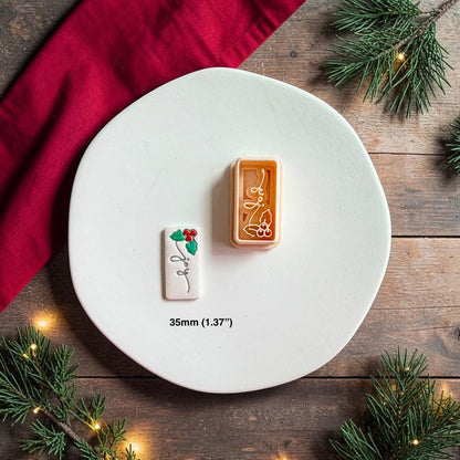 Joy and holly polymer clay cutter beside the finished white clay piece on a white plate, surrounded by red holiday fabric, evergreen branches, and warm lights, with text showing the 35mm or 1.37 inch size.