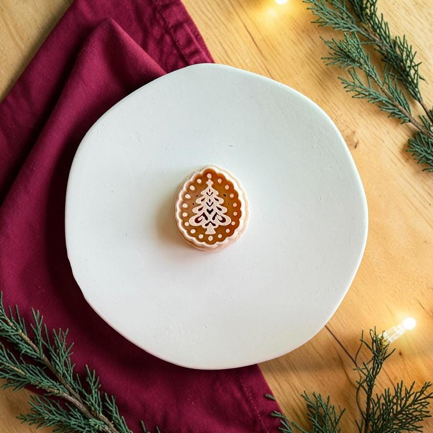 Christmas tree polymer clay cutter centered on a white plate, showing a scalloped oval border, dotted interior frame, and decorative layered pine tree design.
