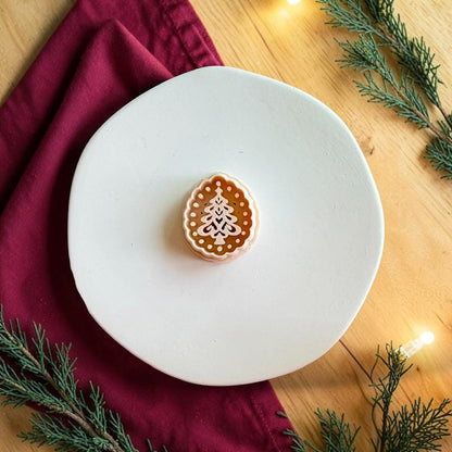 Christmas tree polymer clay cutter centered on a white plate, showing a scalloped oval border, dotted interior frame, and decorative layered pine tree design.