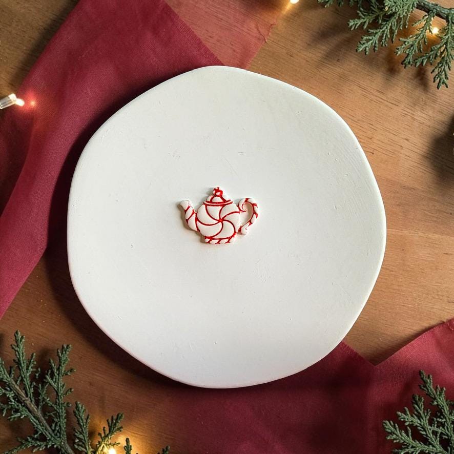 Finished white peppermint teapot clay piece centered on a white plate, featuring bright red swirl stripes across the body, handle, spout, lid, and base against a festive holiday background.