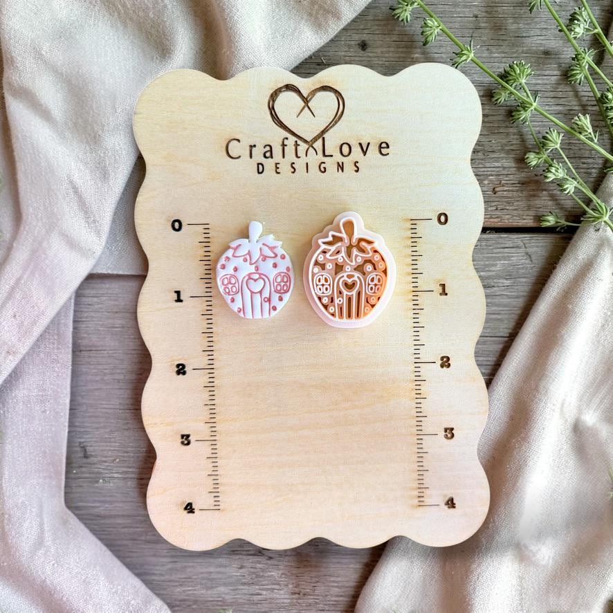 Finished white and pink strawberry house clay piece displayed beside the matching orange design on a Craft Love Designs wooden measurement board.
