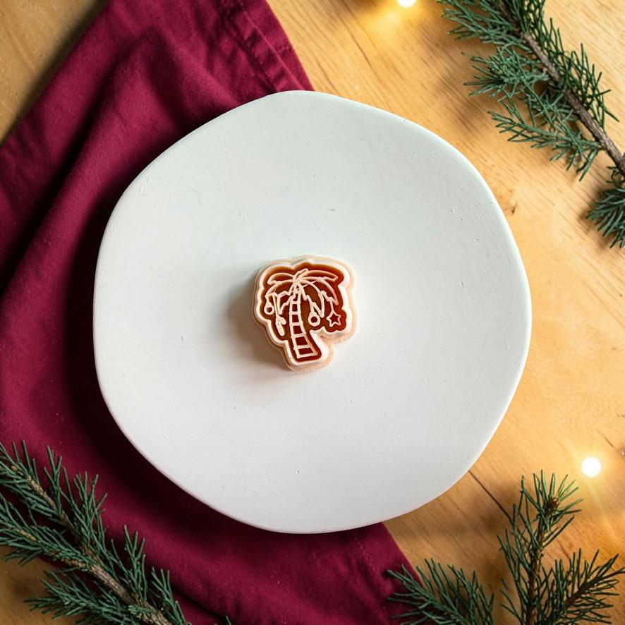 Christmas palm tree polymer clay cutter centered on a white plate, showing a curved trunk, palm leaves, and hanging ornament details against a festive red and evergreen background.