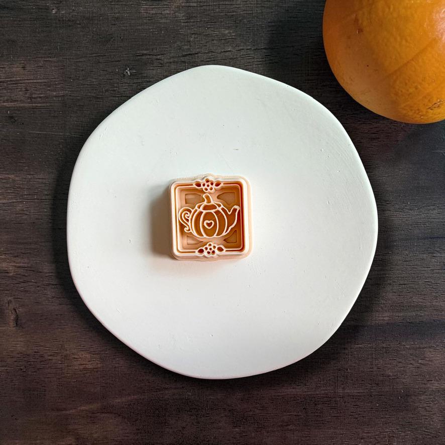 Pumpkin teapot polymer clay cutter centered on a white plate, showing a pumpkin-shaped teapot with curved sections, handle, spout, heart detail, and decorative square frame.