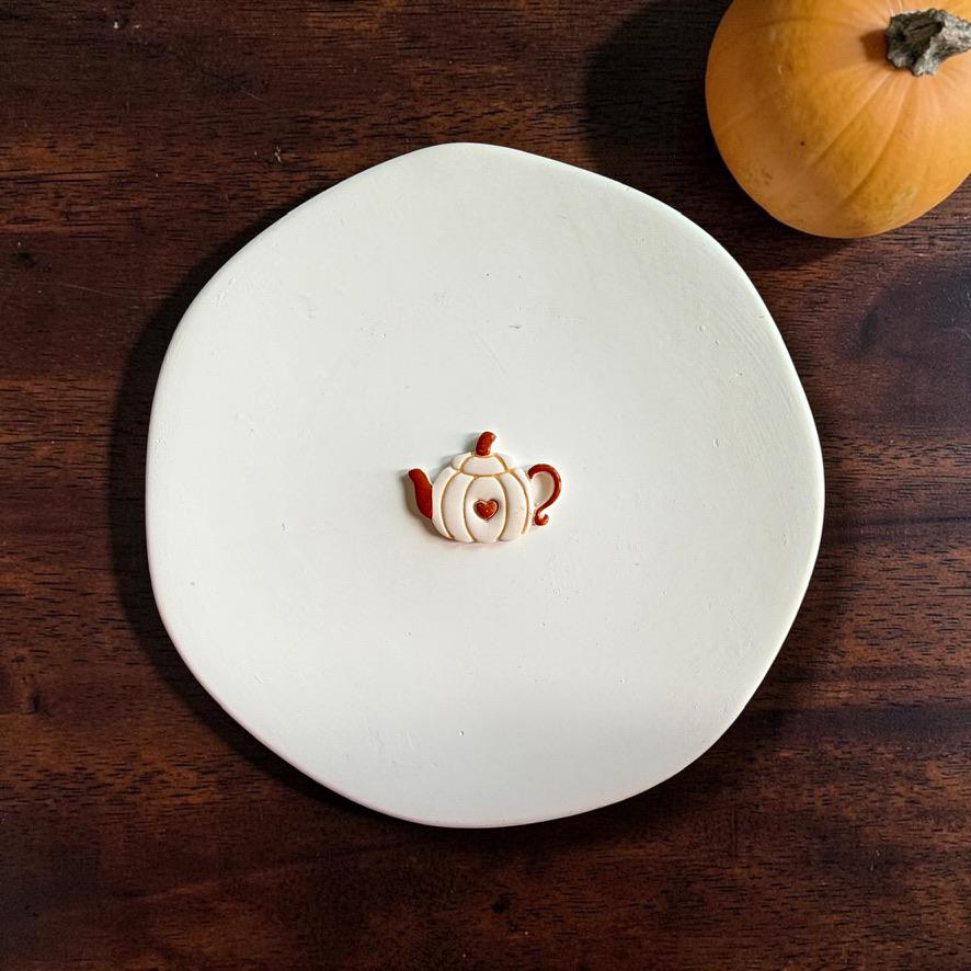 Finished pumpkin teapot clay piece centered on a white plate, showing the white pumpkin-shaped teapot with orange handle, spout, stem lid, and a small heart detail in the middle.
