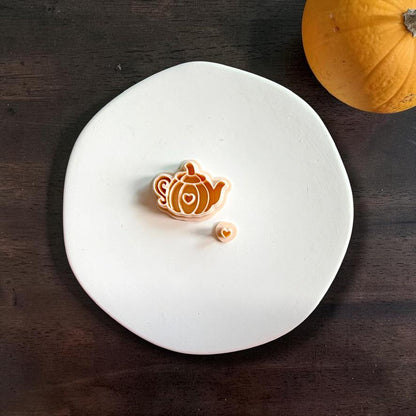 Pumpkin teapot polymer clay cutter centered on a white plate with a small heart-shaped piece next to it, photographed on a dark wood background with a pumpkin in the top corner.