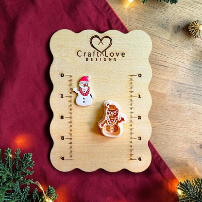 Finished tropical snowman clay piece beside the matching polymer clay cutter on a wooden Craft Love Designs sizing board, with red fabric, greenery, and holiday lights in the background.