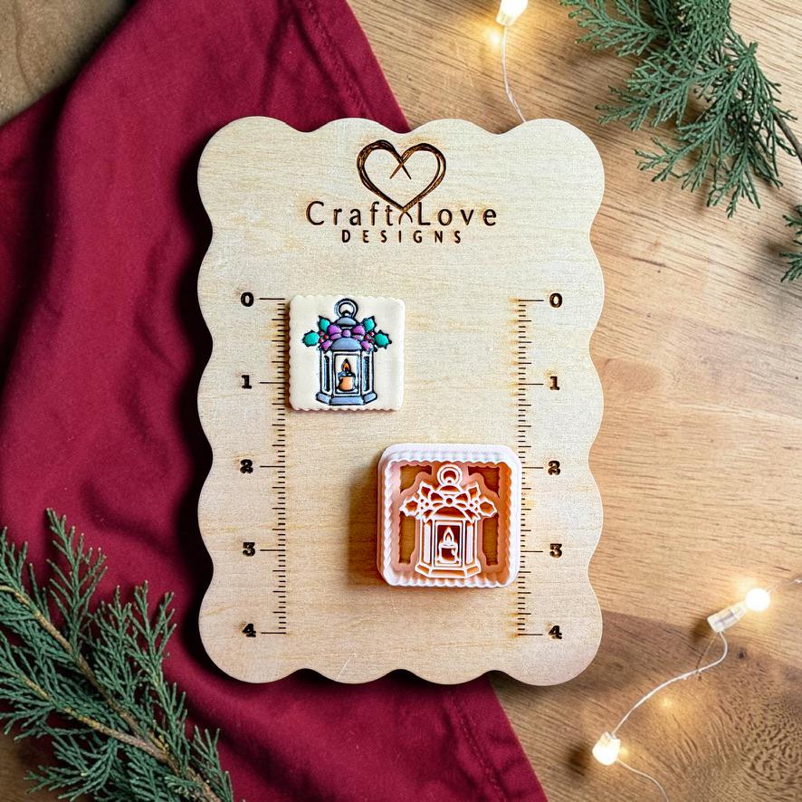 Finished square Christmas lantern clay piece beside the matching polymer clay cutter on a wooden Craft Love Designs sizing board, showing a lantern with a candle, purple bow, green holly leaves, and berry accents.