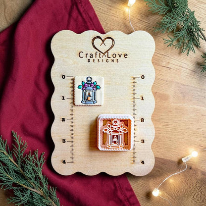 Finished square Christmas lantern clay piece beside the matching polymer clay cutter on a wooden Craft Love Designs sizing board, showing a lantern with a candle, purple bow, green holly leaves, and berry accents.