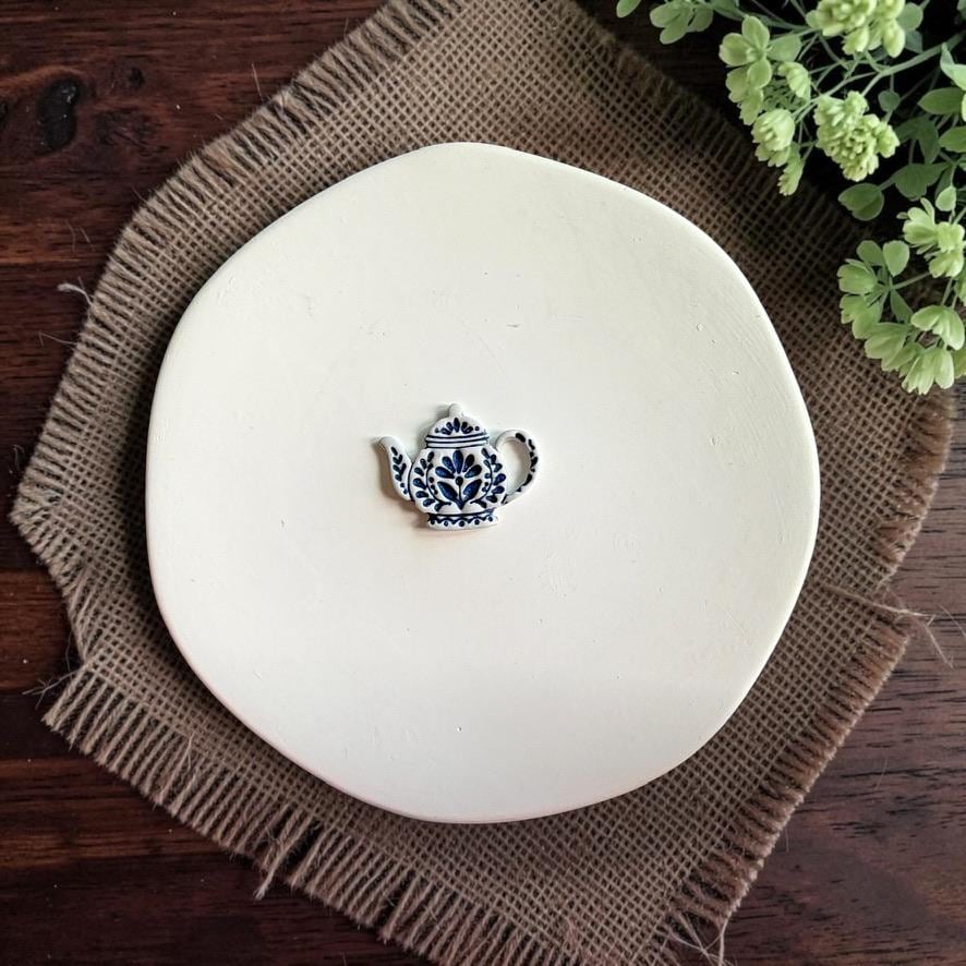Finished folk teapot clay piece centered on a white plate, showing the blue-and-white decorative floral design.