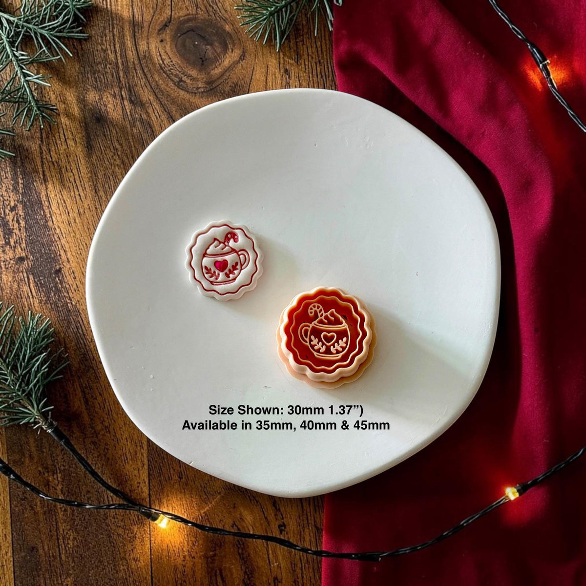 Finished white and red hot cocoa clay piece displayed beside the matching orange design on a white plate with Christmas greenery, red fabric, and warm string lights. Text notes availability in 35mm, 40mm, and 45mm.