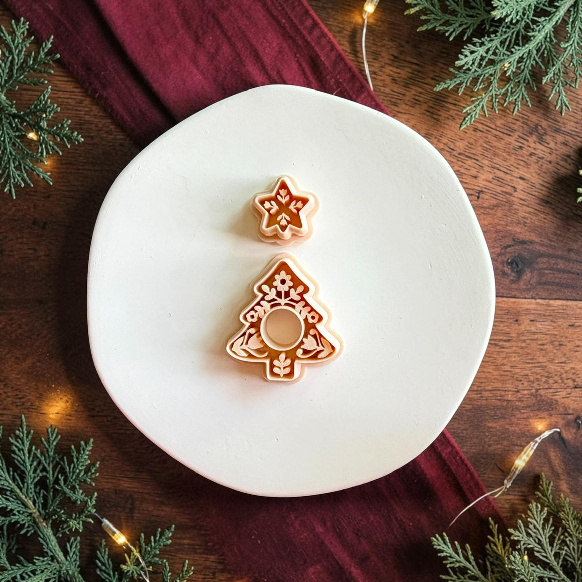 Two-part Christmas tree cutter set on a white plate, featuring a small floral star and larger folk art tree with botanical details and a circular cutout for adding a hanging charm.
