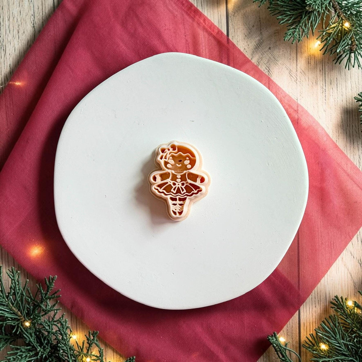 Gingerbread ballerina polymer clay cutter centered on a large white round plate with red holiday fabric, evergreen branches, and warm Christmas lights in the background.