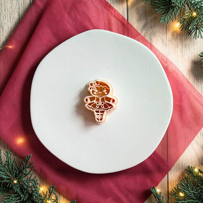 Gingerbread ballerina polymer clay cutter centered on a large white round plate with red holiday fabric, evergreen branches, and warm Christmas lights in the background.