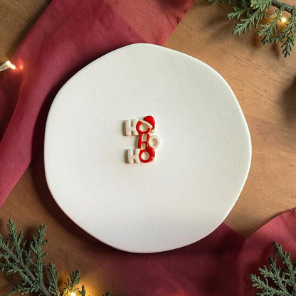 Finished Ho Ho Ho clay piece centered on a white plate, featuring stacked white and red Christmas lettering with a red Santa hat detail.