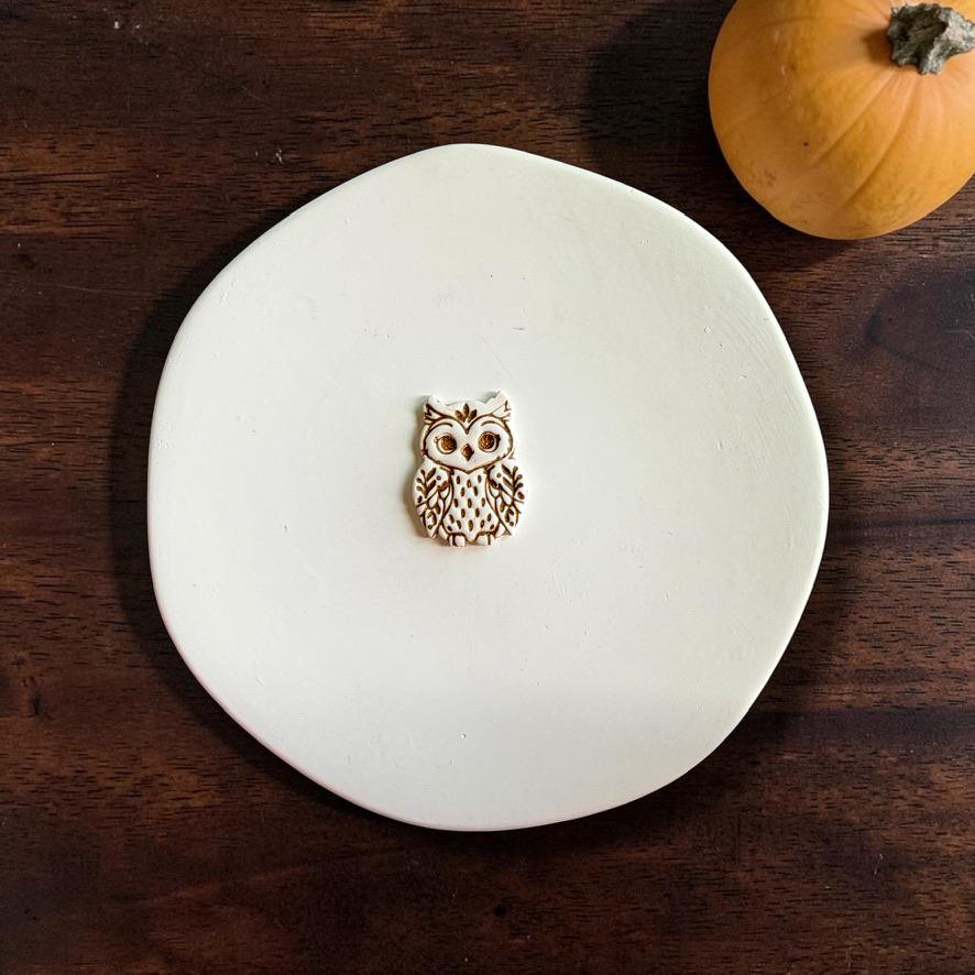 Ornate owl polymer clay cutter centered on a white plate, showing the detailed eyes, wings, feathers, and decorative body pattern, with a small pumpkin in the background.